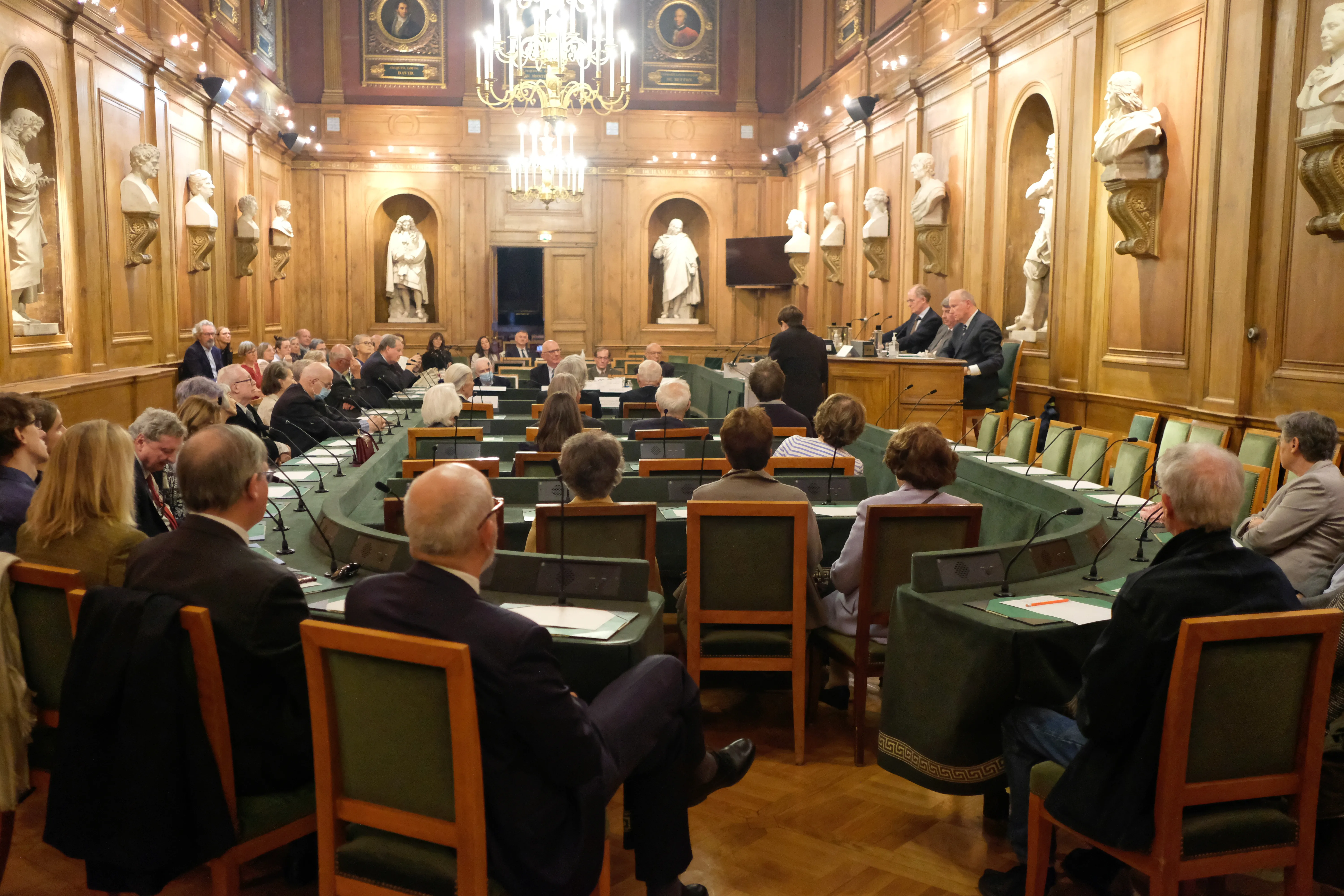 grand pièce de réunion avec des tables vertes, des personnes sont assises et forment un ovale. Elles ont des micros disposés devant elles.